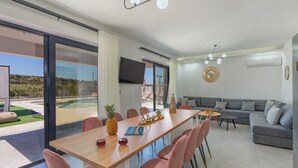 A modern living room with a dining area, a grey sofa, and a pool view. - Luxurious Villa Micha Mare - 14 Bedrooms (Mylopotamos)