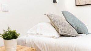 Una cama bien hecha con ropa blanca y dos almohadas estampadas, una mesita de noche de madera con una planta en maceta, y una lámpara fijada a la pared.