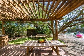 A wooden pergola with a picnic table under a tree, surrounded by greenery. - Koyari Modern Condos 1 (Noord)