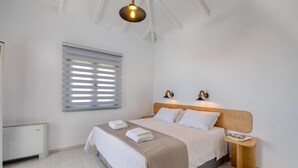 Une chambre à coucher comprenant un lit, des tables de chevet, une fenêtre avec des stores et un luminaire suspendu.