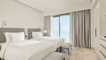 A modern hotel room with two beds, a bench, a large window with curtains, and a view of the sky.
