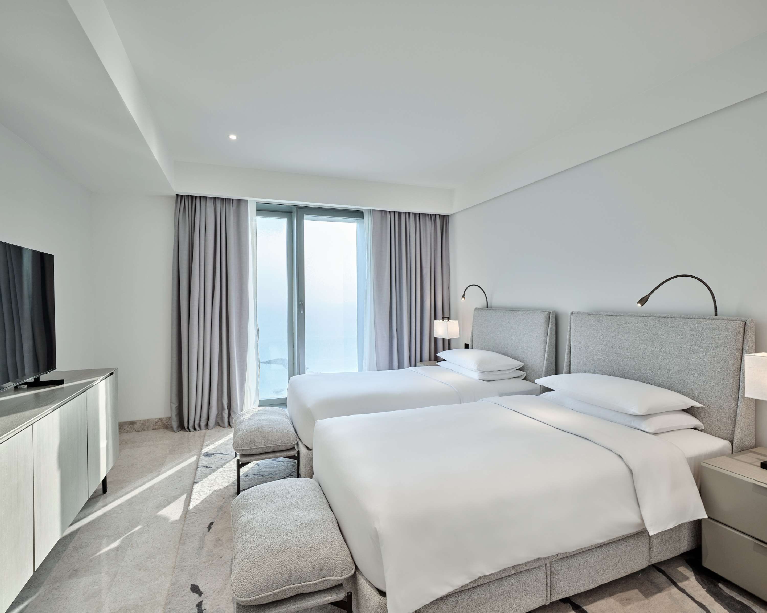 Ein modernes Hotelzimmer mit zwei Betten, einem Flachbildfernseher, einem Schreibtisch und einem großen Fenster mit Vorhängen.