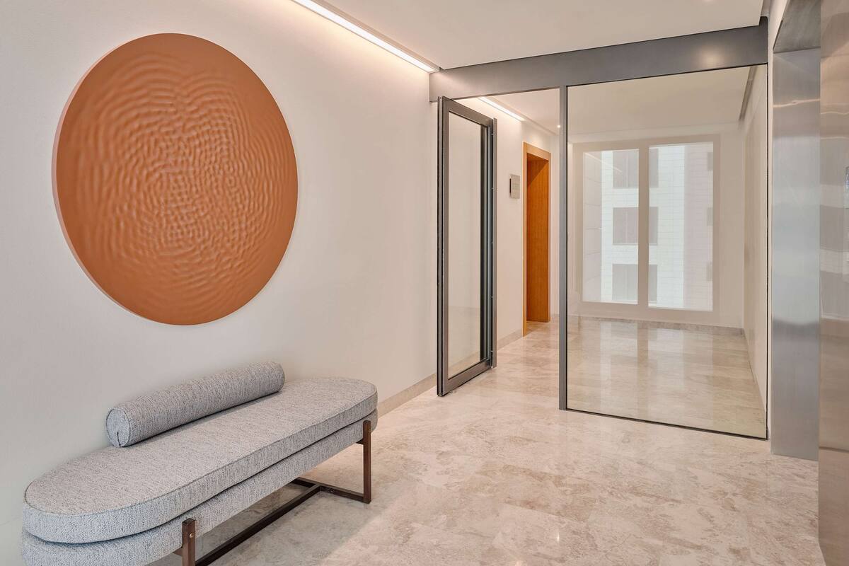 A modern hallway with a bench, a large circular wall art, and a glass door leading to another room.