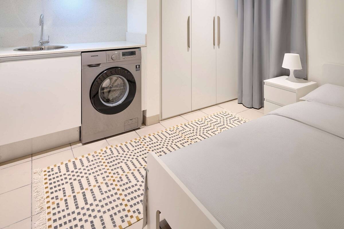 A modern bedroom with a bed, a nightstand with a lamp, a washing machine, and a wardrobe.