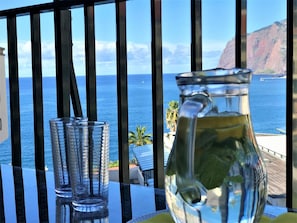 Ein Tisch mit zwei klaren Gläsern und einer Karaffe, mit Blick auf die Küste, das Meer und eine felsige Klippe.