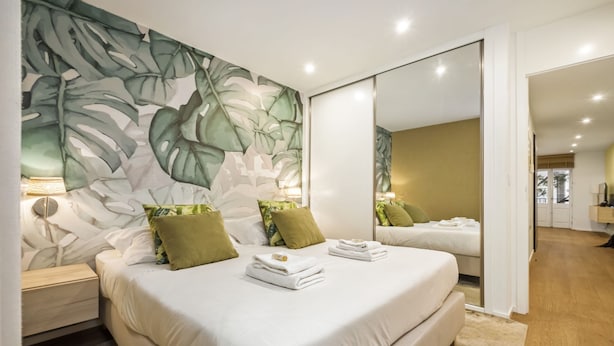 A bedroom with a large bed, a nightstand, a mirror, and a wall with a tropical leaf pattern. - Funchal Tropical - Hurricane Plant by Homie (Funchal)