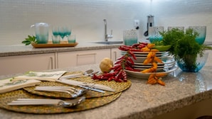 A kitchen counter with a woven placemat, a bowl of red chili peppers, a bunch of fresh herbs, and a stack of plates. - Funchal Tropical - Hurricane Plant by Homie (Funchal)