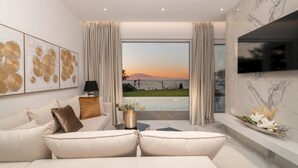 A modern living room with a sofa, a TV mounted on the wall, and a view of the sea at sunset.