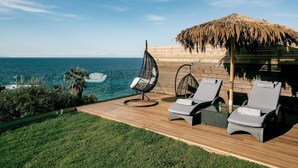 A deck with two lounge chairs, a hanging chair, and a thatched roof structure overlooking the sea.