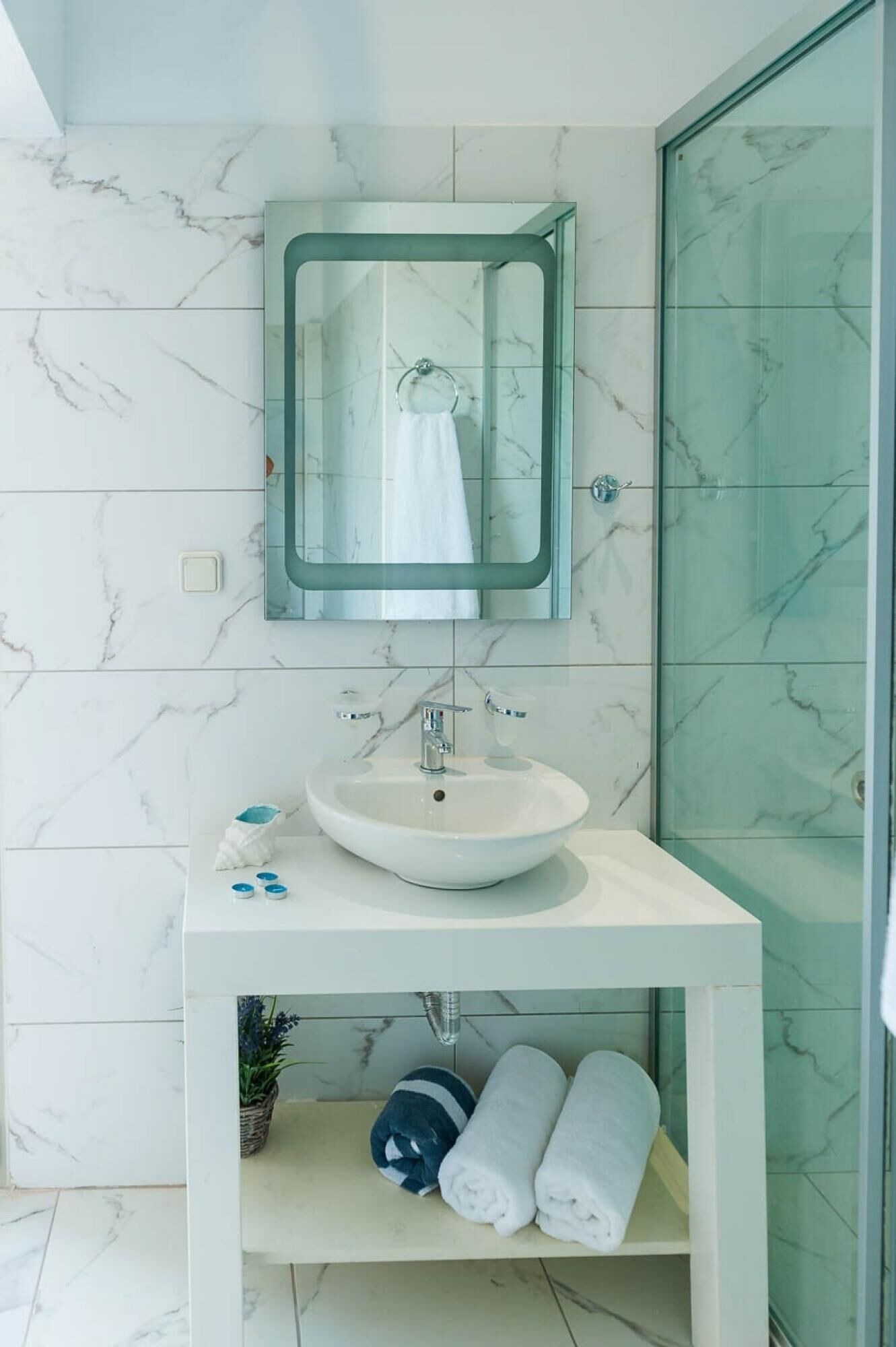 A modern bathroom with a marble-tiled wall, a white pedestal sink, a glass-enclosed mirror, and a glass shower enclosure.