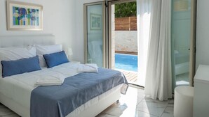 A bedroom with a bed, a nightstand, a painting, and a view of an outdoor swimming pool. - Villa Prima - With Private Heated Pool (Rethymno)