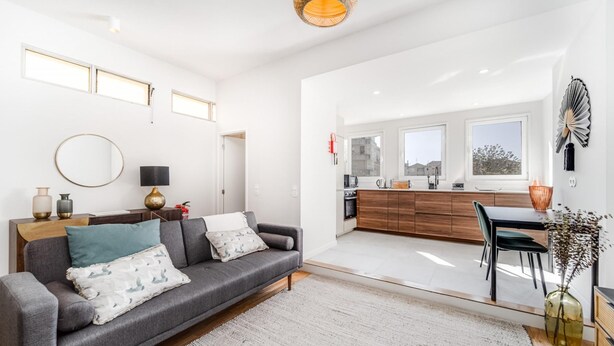 A modern living room with a grey sofa, a coffee table, and a dining area with a table and chairs. There is a kitchen visible through an open door. - Nomad s By Sta Catarina Flats - 3bdr Family (Porto)