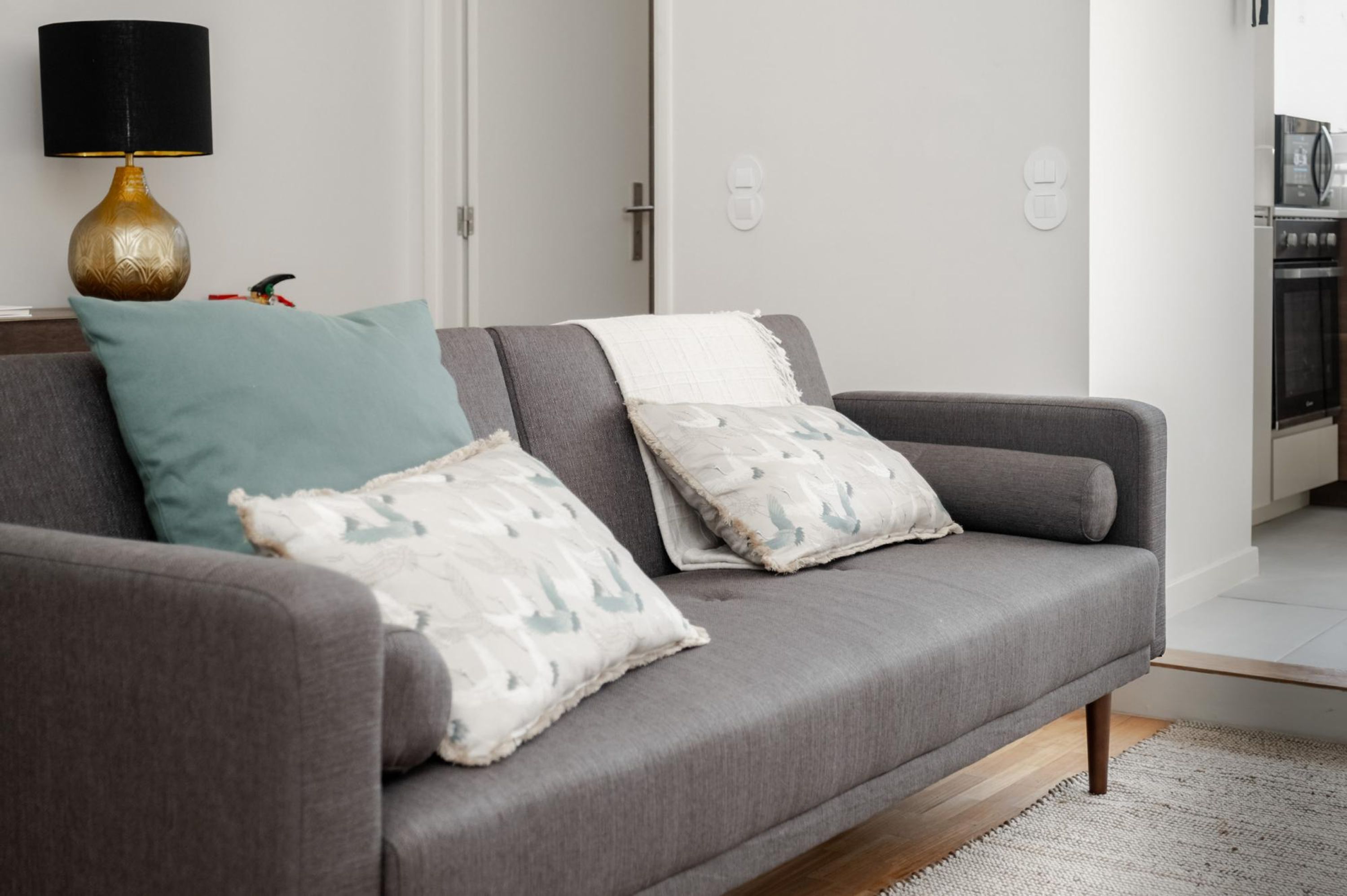 Un salon moderne avec un canapé gris, des coussins à motifs turquoise et blancs, une lampe dorée et une cuisine visible au fond.