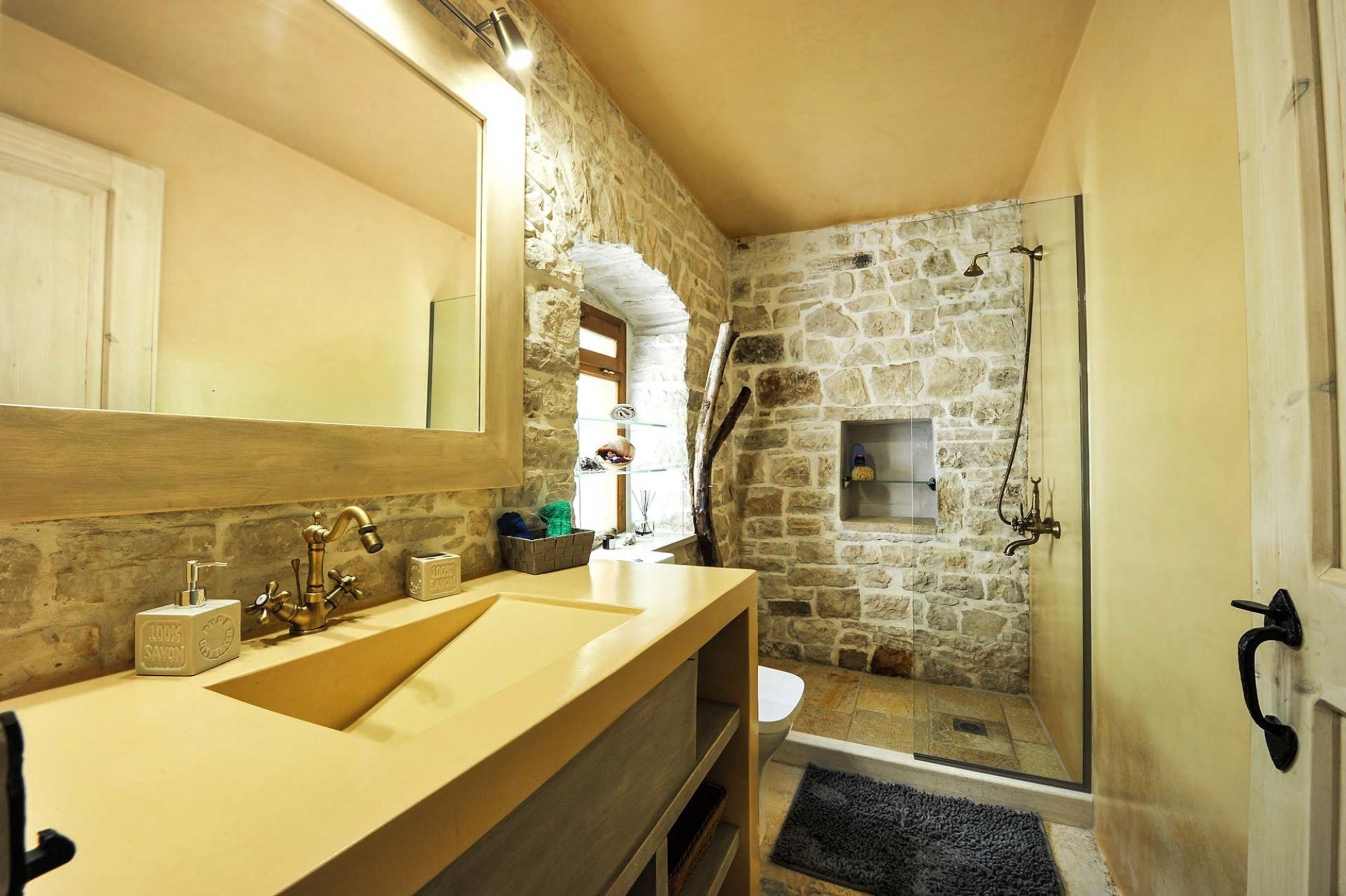 A bathroom with a stone wall, a wooden vanity, a large mirror, and a shower area.