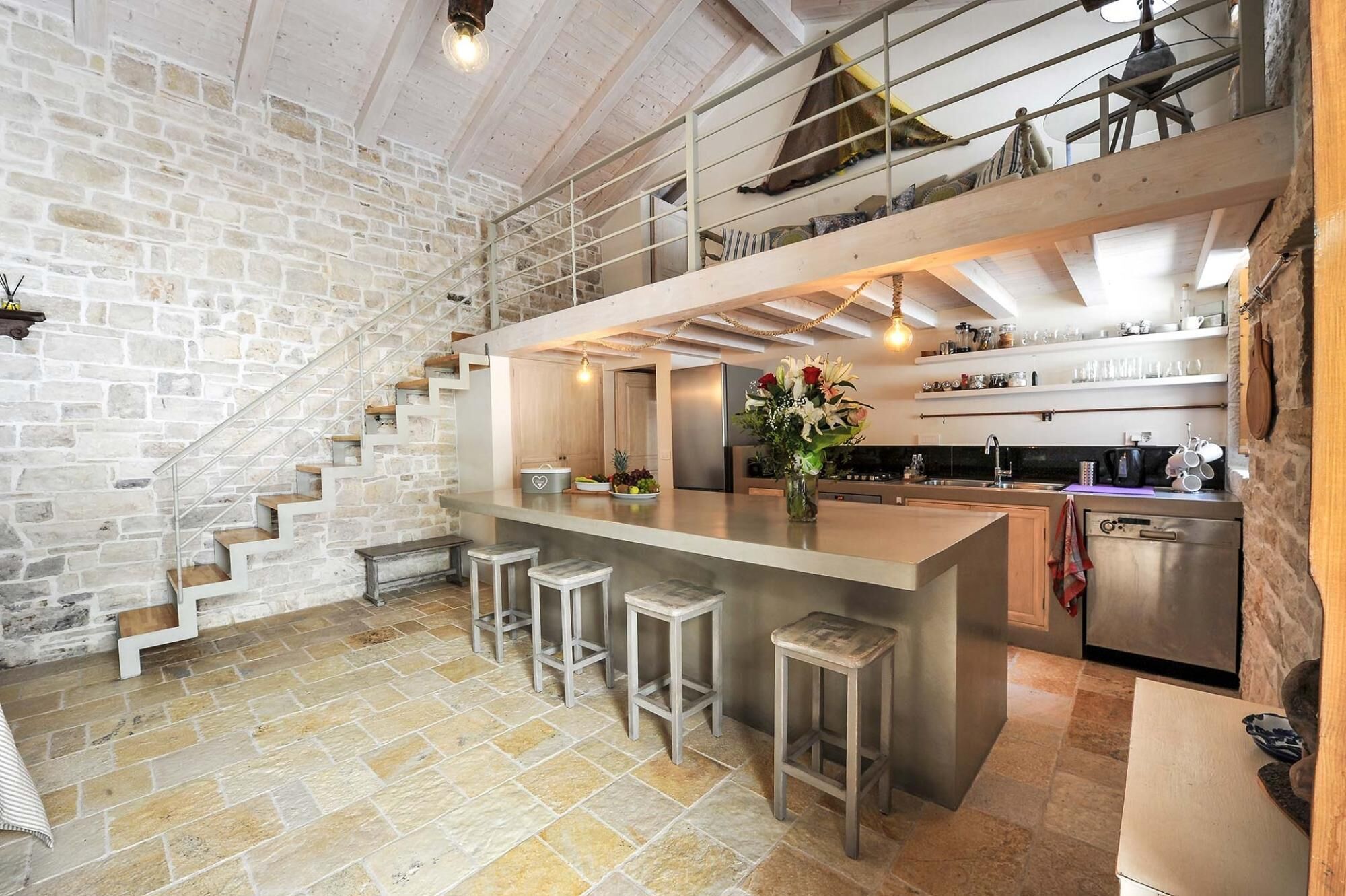 A spacious kitchen with a central island, a stone wall, and a staircase leading to an upper level.
