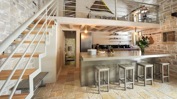 A spacious loft with a high ceiling, exposed wooden beams, a stone wall, a kitchen area with a bar, and a staircase with a metal railing. - Leonora House With Private Pool Close to Gaios (Paxos)