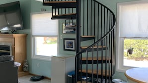 Un espacio habitable compacto con una escalera en espiral, un televisor, una mesita y una ventana con vistas a zonas verdes.
