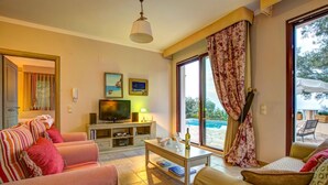 A living room with a sofa, armchair, TV, and a table with a bottle and glasses. - Theodora Villa - 2 BR Tranditional House Close to Gaios Town (Paxos)