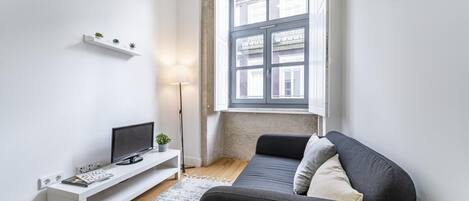 Uma sala de estar moderna com um sofá cinzento, um rack branco para TV, uma planta pequena e uma janela grande com vista para edifícios.