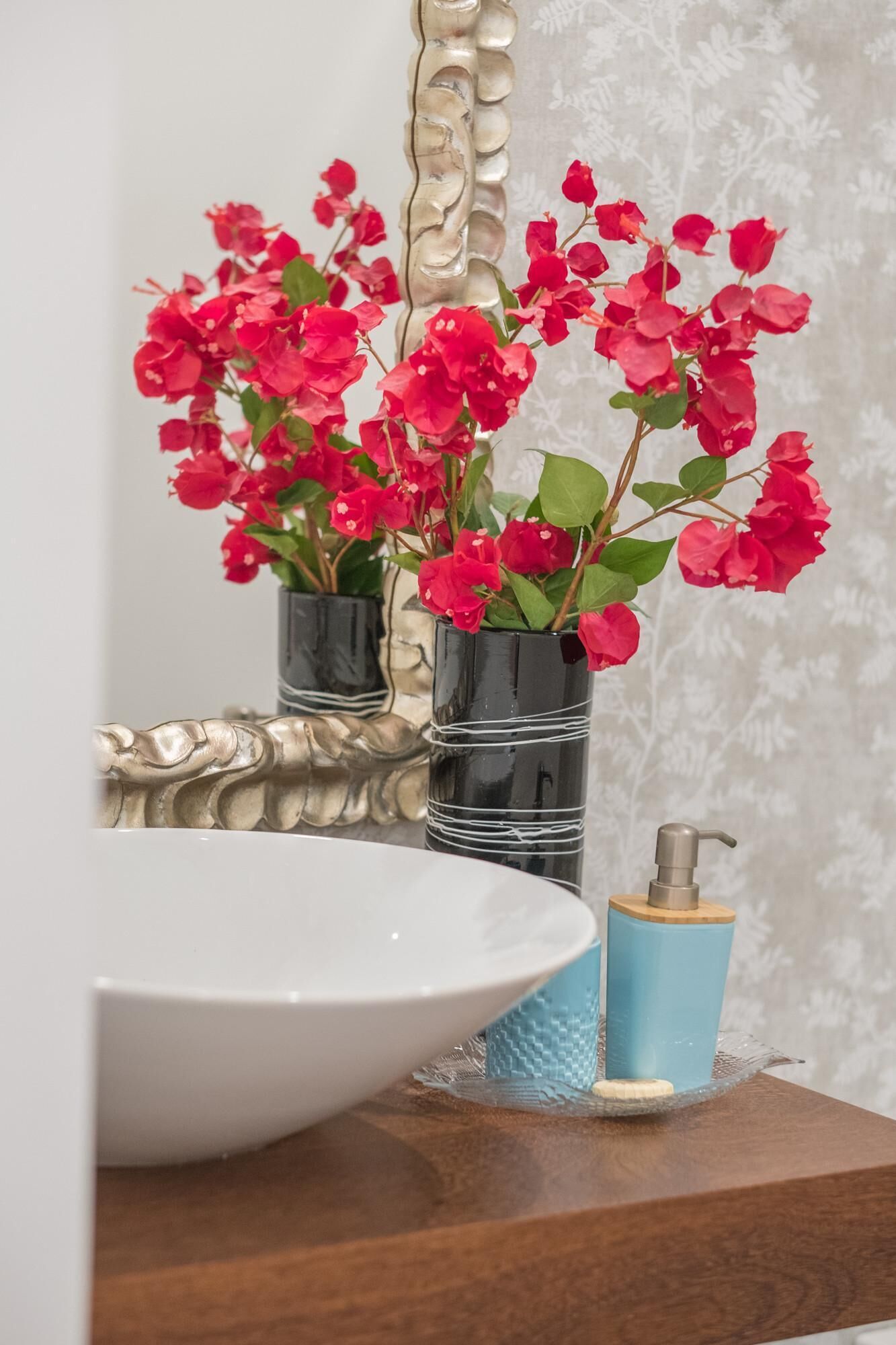 Un tocador de baño con un lavamanos blanco, un dispensador de jabón azul y dos jarrones negros con flores rojas.