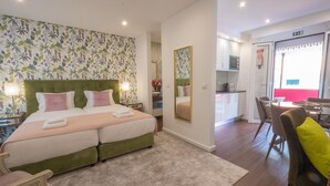 Une chambre à coucher avec un lit double, une tête de lit recouverte d’un tissu vert, un miroir et une petite table de chevet avec une lampe.