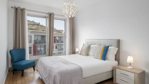 Ein modernes Schlafzimmer mit einem großen Bett, einem Sessel, einem Nachttisch mit Lampe und Blick auf Gebäude durch das Fenster.