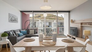 Ein modernes Wohnzimmer mit einer Couch, einem Esstisch und Stühlen. Ein großes Fenster bietet Blick auf Gebäude und Hügel.