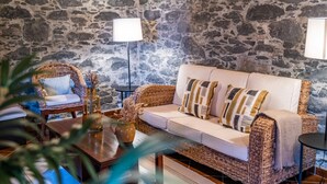 A cozy living room with a stone wall, a wicker sofa with cushions, a wooden coffee table, and a floor lamp. - Rainbow Cottage by Homie (Calheta)
