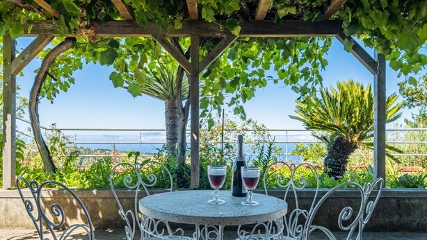 A round table with metal chairs under a pergola with green vines, overlooking a scenic view. - Rainbow Cottage by Homie (Calheta)