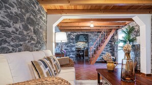 A cozy living room with a stone wall, a wooden ceiling, and a staircase leading to a dining area. - Rainbow Cottage by Homie (Calheta)