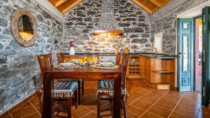 A rustic stone interior with a dining table set for four, wooden chairs, a stone wall, and a kitchen area with a sink and cabinets. - Rainbow Cottage by Homie (Calheta)