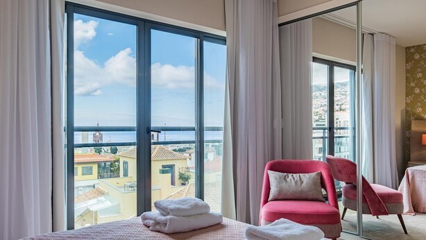 Ein Hotelzimmer mit großem Fenster, einem Bett mit ordentlich gefalteten Handtüchern, einem roten Sessel und Blick auf die Stadtlandschaft.