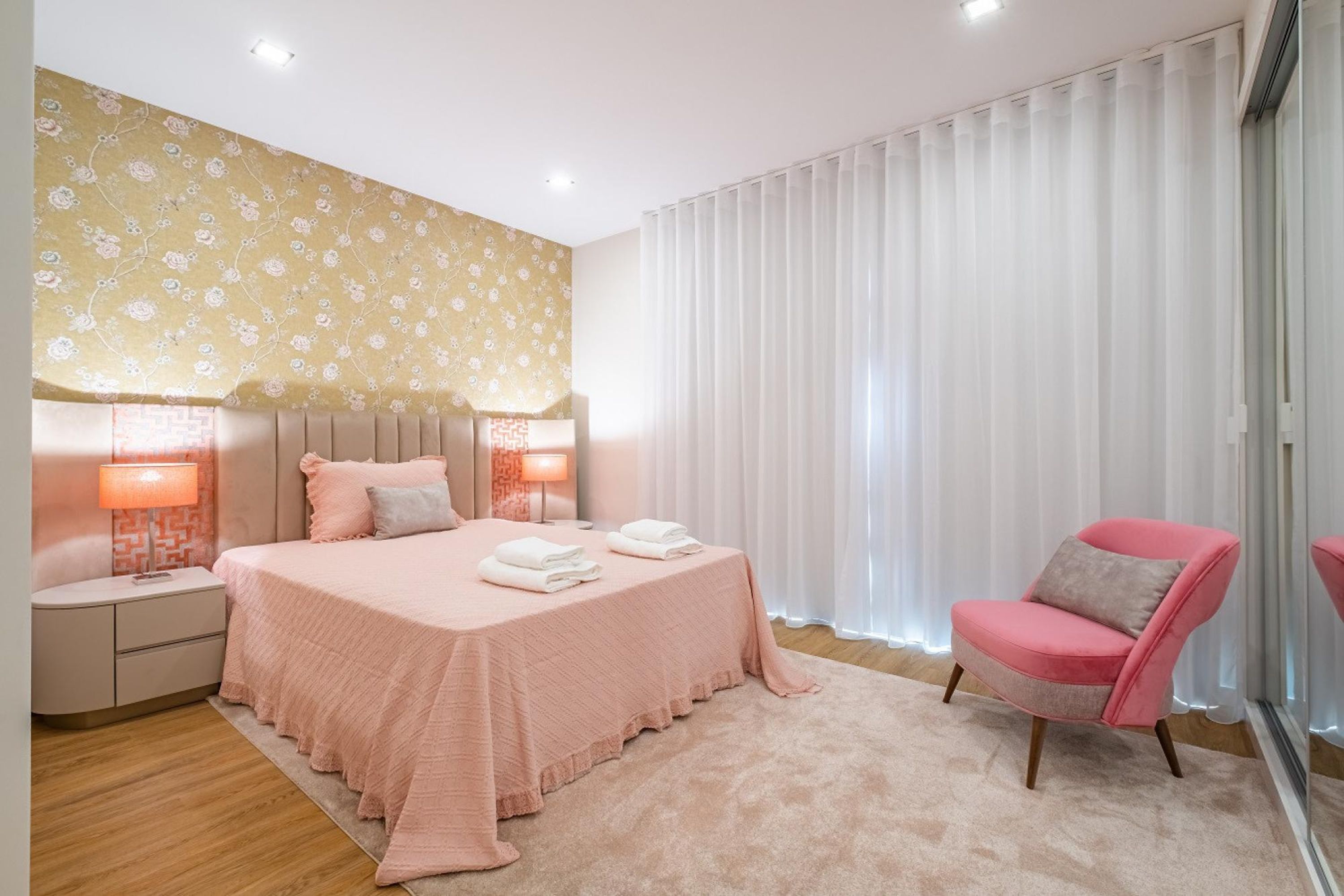 Een slaapkamer met een bed, nachtkastjes, een stoel en een groot raam met gordijnen.