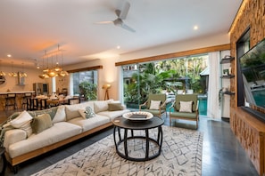Relax in this spacious living area with a large sofa, coffee table, and dining table, offering a view of the lush garden and pool.