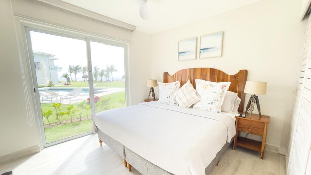 A bedroom with a large bed, wooden headboard, two bedside tables with lamps, and a view of a pool and greenery through a sliding glass door. - Residences C2-pbb in Chame (Punta Chame)