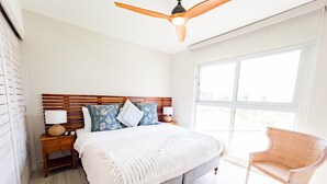 A bedroom with a wooden headboard, a large bed with a white blanket, a nightstand with a lamp, a chair, and a ceiling fan. - Residences C4-1c in Chame (Punta Chame)