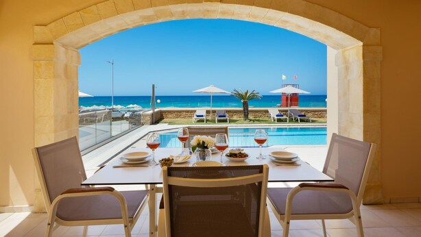 Een eettafel met twee stoelen, met uitzicht op een zwembad en een strandgedeelte met parasols en ligbedden.
