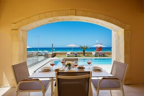 A dining table with two chairs, a view of a pool, and a beach area with umbrellas and lounge chairs. - Beachfront Villa Avra With Private Swimming Pool (Rethymno)