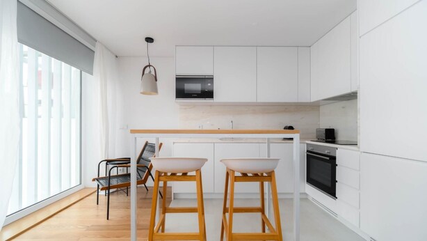Una cocina moderna con armarios blancos, encimera de madera y horno empotrado. Hay dos taburetes de madera y una lámpara colgante.