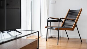 Una sala de estar moderna con piso de madera, una mesa de centro de vidrio, una silla negra y de madera, y un televisor sobre un mueble.