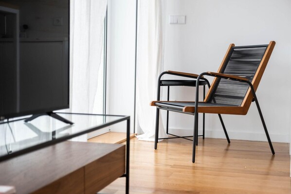Een moderne woonkamer met een houten vloer, een glazen salontafel, een zwarte en houten stoel, en een televisietoestel op een meubel.
