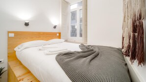 A bedroom with a wooden headboard, white bedding, a gray blanket, and a macrame wall hanging.