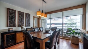 A modern dining area with a wooden table and leather chairs, large windows with a view of buildings, and hanging pendant lights.