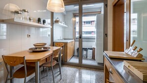 A modern kitchen with a wooden table, chairs, a hanging light fixture, and a view through a glass door to the outside.