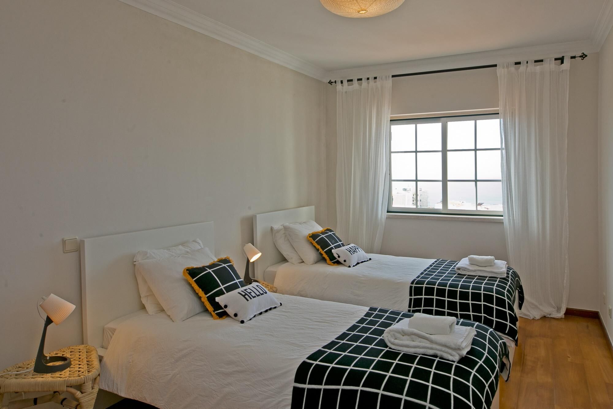 A hotel room with two beds, white bedding, a wicker chair, a bedside table, a lamp, and a window with curtains.