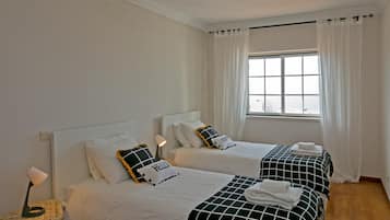A hotel room with two beds, white bedding, a wicker chair, a bedside table, a lamp, and a window with curtains.