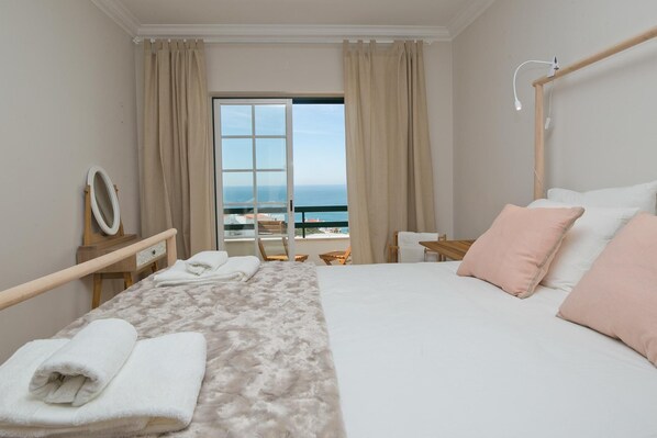 A hotel room with a bed, a bedside table with a lamp, a mirror, and a view of the sea through a large window.