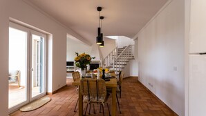 A spacious dining area with a wooden table, wicker chairs, and a hanging light fixture. A staircase is visible in the background leading to an upper level. - Villa Octopus s Garden by Acasadascasas (Mafra)