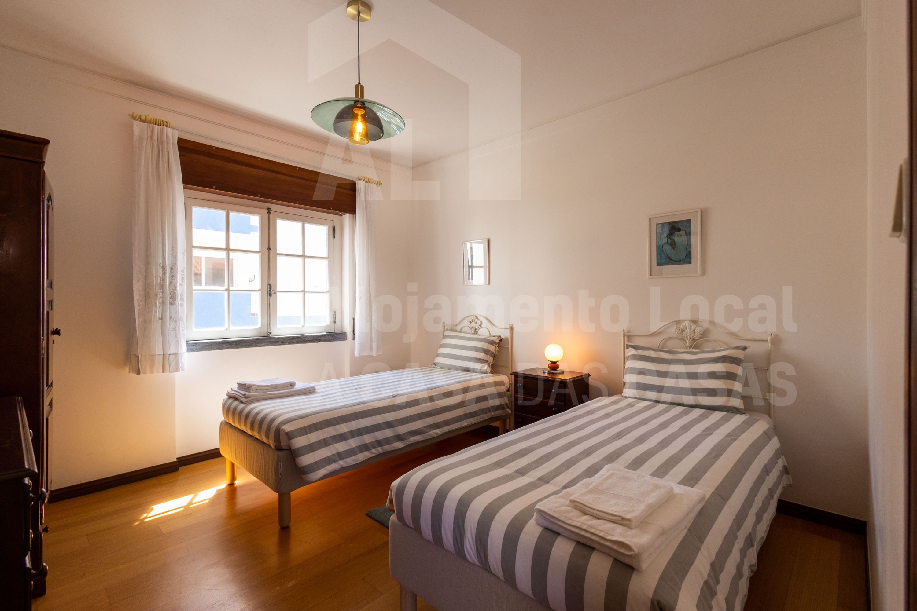 Una camera da letto con due letti singoli, una finestra con tende, un comodino con una lampada e un quadro appeso al muro.