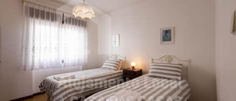 Une chambre avec deux lits simples, une table de chevet avec une lampe, une fenêtre avec des rideaux légers et un tableau au mur.